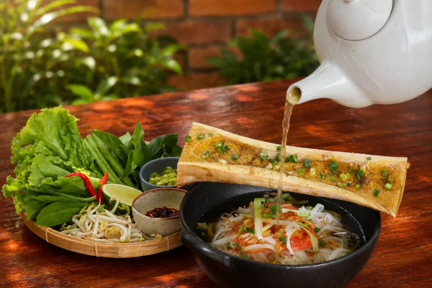 Broth poured over a noodle soup topped with roasted bone marrow, served with fresh herbs and greens